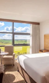 Bright hotel room with a large bed, armchairs, table, and expansive countryside view.