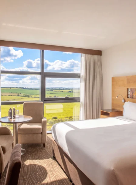 Bright hotel room with a large bed, armchairs, table, and expansive countryside view.