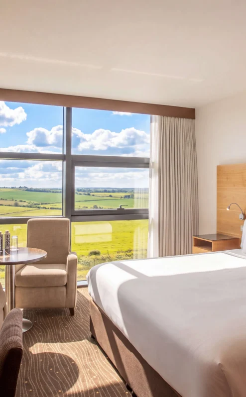 Bright hotel room with a large bed, armchairs, table, and expansive countryside view.