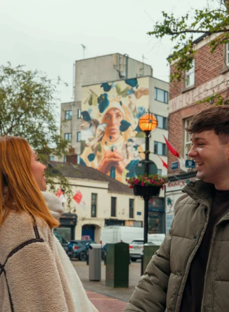 A couple smiles and enjoys a stroll outside The Gateway Hotel, Dundalk, with street art and trees nearby.