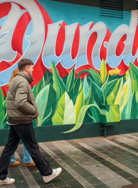 A couple walks past a colourful mural with "Dundalk" in vibrant letters, near The Gateway Hotel.