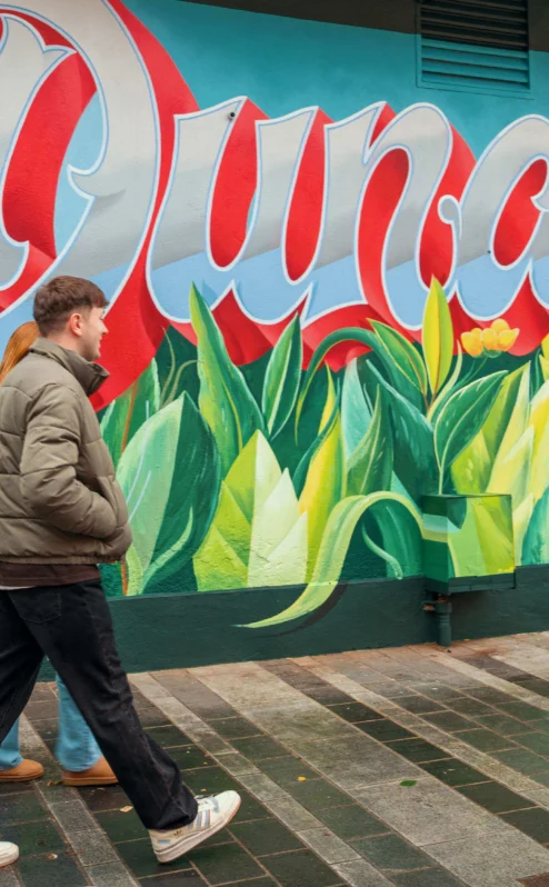A couple walks past a colourful mural with "Dundalk" in vibrant letters, near The Gateway Hotel.