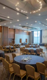 Spacious hotel event hall with round tables set for a gathering, warm lighting, and large windows.