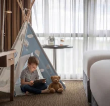 A child reads on a tablet beside a teddy bear in a cosy hotel room with a teepee tent.
