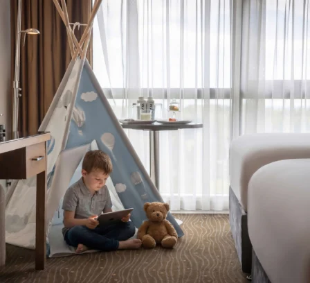 A child reads on a tablet beside a teddy bear in a cosy hotel room with a teepee tent.