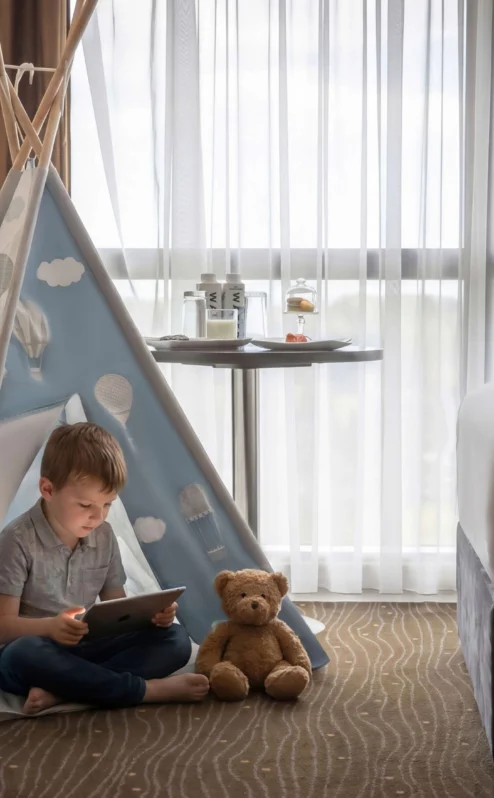 A child reads on a tablet beside a teddy bear in a cosy hotel room with a teepee tent.