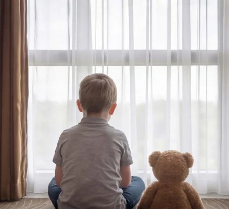 Young boy with teddy bear gazing out bright hotel window.