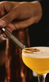 A hand garnishes a cocktail with a dried orange slice, set in a cosy bar at The Gateway Hotel.