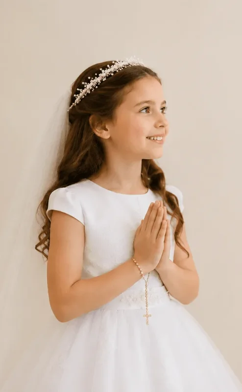 Young girl in white dress with veil, smiling and holding rosary, against neutral background.